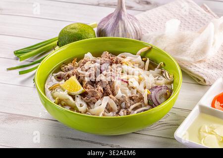 Eine leuchtend grüne Schüssel mit traditioneller vietnamesischer Rindfleischnudelsuppe Pho bo, mit Reisnudeln, Sojasprossen und Zwiebeln. Serviert mit frischer Limette, Chili, an Stockfoto
