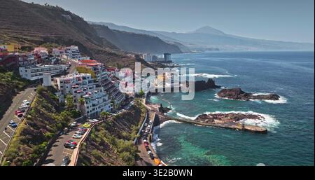 Teneriffa: Drohnenflug über farbenfrohe Hotelresort-Gebäude mit Küstenstädten auf einem Hügel, Pier, tiefblauem Atlantik, mit dem fernen Vulkan Pico del Teide im Hintergrund Stockfoto