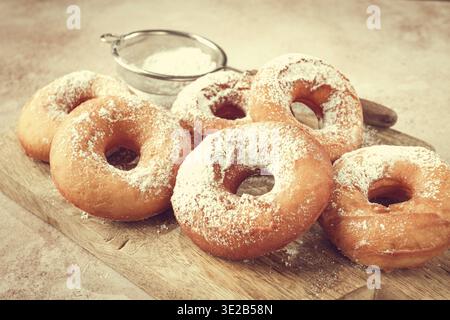 Auf einem Holzbrett befinden sich mehrere frisch zubereitete Donuts. Die Donuts sind rund und haben Puderzucker drauf. Ein kleines Sieb für zusätzlichen Zucker ist in der Nähe Stockfoto