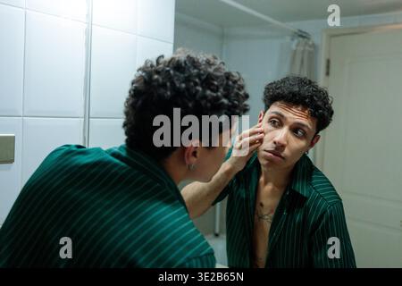 Junger hispanischer Mann, der das Gesicht im Badezimmerspiegel überprüft Stockfoto