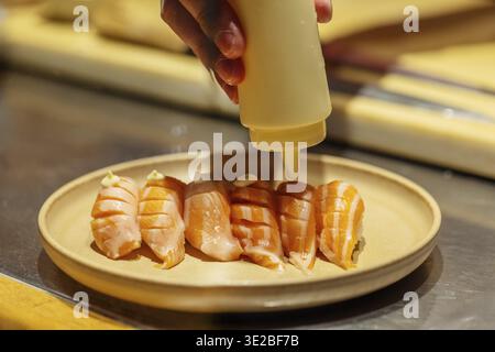 Der Sushi-Koch fügt gekonnt eine Sauce zu einem Teller Lachs-Sushi hinzu und zeigt kulinarische Kunst und Liebe zum Detail in einer lebendigen Küche Stockfoto
