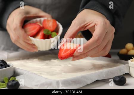 Joghurtrinde herstellen. Frau, die die Hälfte der Erdbeere auf das Geschirr mit Joghurt auf den weißen Tisch legt, Nahaufnahme Stockfoto