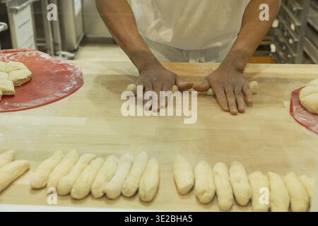 Ein erfahrener Bäcker formt auf einer Holzarbeitsfläche Teig zu Rollen, umgeben von Backwerkzeugen und Zutaten, die kulinarische Handwerkskunst zeigen Stockfoto