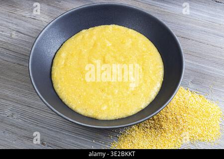 Gekochter Maisbrei in einer grauen Schüssel und eine Handvoll Maismehl auf einem grauen Holztisch Stockfoto