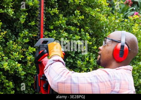 Afroamerikaner schneidet Hecke mit rotem Trimmer im Hof, trägt Handschuhe, Brille, Kopierraum. Gartenbau, Outdoor, Gartenbau, Landschaftsgestaltung, kabellos, ej Stockfoto
