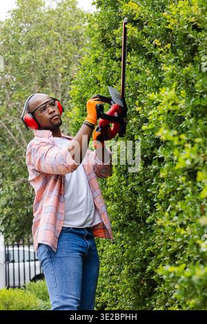 Afroamerikaner, der große grüne Hecke im Vorhof mit rotem Trimmer trimmt und Sicherheitsausrüstung trägt. Im Freien, Landschaftsgestaltung, Gartenbau, Wohnbereich, Garten Stockfoto