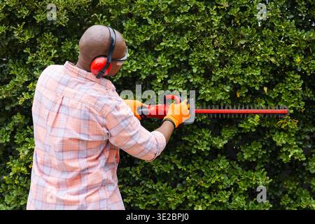 Erwachsener afrikanischer Mann schneidet Hecke im Hof mit roter Heckenschere, trägt orangefarbene Handschuhe, Kopierraum. Gärtner, Beschneiden, Laub, Garten, draußen, v Stockfoto