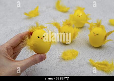 Süße gelbe Ostereier in Form von Küken, umgeben von Federn auf grauem Hintergrund. Die Hand einer Frau demonstriert ein Handwerk, das aus einem Ostern hergestellt wurde Stockfoto