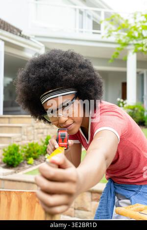 Afrikanische Frau mit Schutzbrille, die Holzdiele im Vorhof mit gelbem Klebeband misst. Holzbearbeitung, diy, Outdoor, Vorort, Werkzeuge, Klemme, Veranda Stockfoto