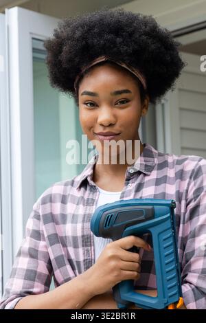 Frau in 20ern, die in der Tür steht und einen blauen kabellosen Power-Nagler hält, mit kariertem Hemd. Werkzeug, Handgerät, heimwerker, Hausbesitzer, Tageslicht, Schwellenwert, por Stockfoto