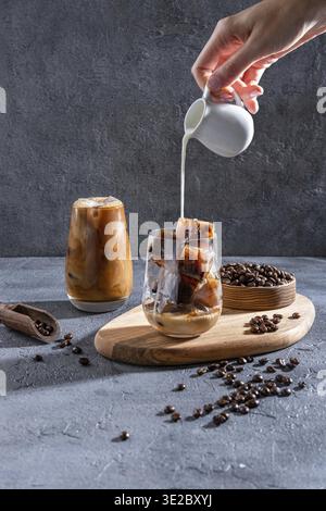 Milch wird auf einem dunklen Tisch in Eiskaffee gegossen. Eiskaffee in einem großen Glas mit Sahne und Kaffeebohnen. Kaltes Kaffeegetränk auf einem dunklen Ba Stockfoto
