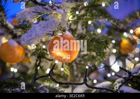 Orangefarbene Ornamente mit Schnee auf einem Weihnachtsbaum, beleuchtet von warmweißen Lichtern in festlicher Winteratmosphäre Stockfoto