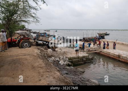 Satkhira, Bangladesch - 25. April 2025: Ansicht von Arbeitern, die Bausteine von einem mit einem Traktor gezogenen Wagen auf einen teilweise gebauten Pier abladen Stockfoto