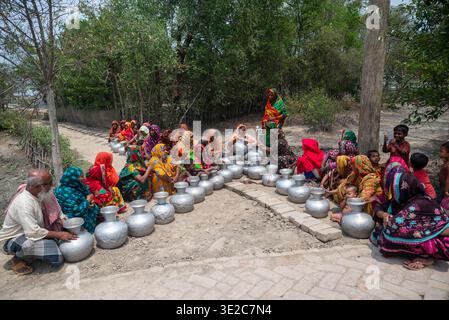 Satkhira, Bangladesch - 25. April 2025: Blick auf Frauen in leuchtender Saris-Warteschlange mit glänzenden Metalltöpfen, die den grellen Glanz der Sonne auf dem park widerspiegeln Stockfoto