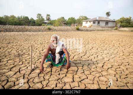 Satkhira, Bangladesch - 25. April 2025: Blick auf einen verwitterten Mann, der sich in einer gerösteten, zerrissenen Erde hockte, eine krasse Landschaft, die sich bis zu einem entfernten Haus erstreckt. Stockfoto
