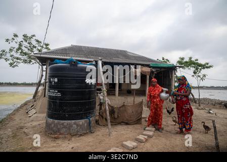 Satkhira, Bangladesch - 25. April 2025: Blick auf ein bescheidenes Haus, in dem zwei Frauen in leuchtenden roten und orangen Saris Wasser unter einem bewölkten Himmel sammeln. Stockfoto