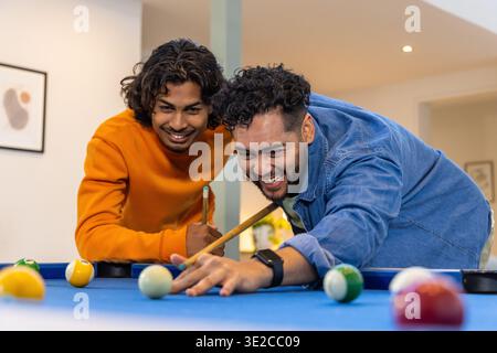 Indische Freunde in Denim-Hemd und orangefarbenem Sweatshirt, die auf den Billardtisch aus blauem Filz zielen. Billard, Duo, Kameradschaft, Heim, Gameroom, modern Stockfoto