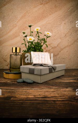 Elegantes Muttertag-Arrangement mit Parfümflasche, verpacktem Geschenk, Gänseblümchen und handgeschriebener Karte auf rustikalem Holz, entworfen für Schönheit und Feier c Stockfoto