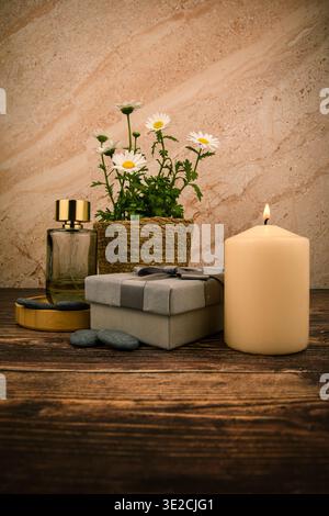 Beleuchtete Kerze, Gänseblümchen, Parfümflasche und Geschenkverpackung auf rustikalem Holz, ein gemütliches Stillleben im Spa zum Muttertag mit warmem Licht und elegantem Design. Stockfoto