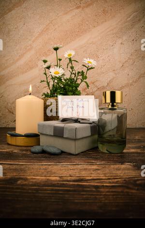 Mutter-Tag-Spa-Geschenkszene mit beleuchteter Kerze, Parfümflasche, Gänseblümchen, handgeschriebener Karte und gewickeltem Geschenk auf rustikalem Holz in warmem, elegantem Stil. Stockfoto