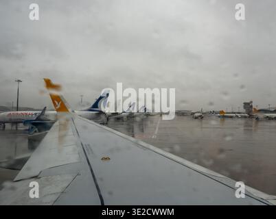 Blick aus einem Flugzeugfenster auf mehrere geparkte Passagierjets auf einem regnerischen Flughafenvorfeld mit nassem reflektierendem, asphaltierendem, grauem Himmel Stockfoto