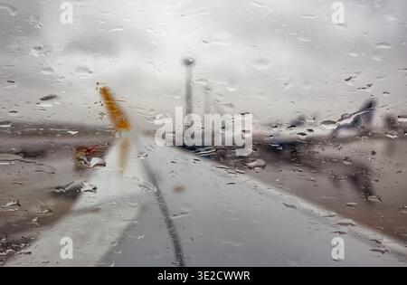 Regentropfen auf einem Flugzeugfenster verdecken die verschwommene Start- und Landebahn des Flughafens und geparkte Jets bei bewölktem Wetter und schaffen so einen stimmungsvollen Reise- und Luftfahrthintergrund Stockfoto