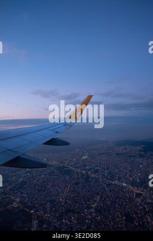 Die Luftaufnahme von einem Passagierfenster zeigt einen Flugzeugflügel über einer dichten Stadt in der Abenddämmerung mit beleuchteten Straßen, Wolken und tiefblauer Dämmerung, die eine Reise A schaffen Stockfoto