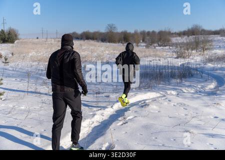 Paare in schwarzem Gear auf schneebedeckten Pfaden auf offenem Feld. Heller Wintertag mit klarem blauem Himmel. Outdoor-Training, gesunder Lebensstil und kaltes Wetter Stockfoto