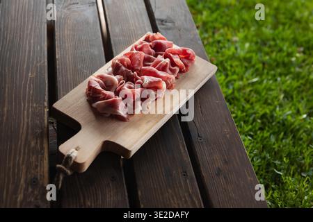 Scheiben italienischer geräucherter Schinken auf einem hellen Holzbrett, dunkler Tisch im Freien, grüner Grashintergrund. Natürliches Sommerlicht, authentischer italienischer Bauernhof A Stockfoto