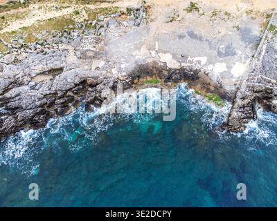 Top-down-Luftaufnahme von Tonnara del Secco in San Vito Lo Capo. Das tiefblaue Meer verfärbt sich in türkisfarbenem Blau an den zerklüfteten Klippen, neben unbefestigten Pfaden und Stockfoto