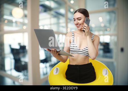 Glückliche Frau im Badeanzug, die im Büro den aufblasbaren Ring hält. Zeit für Urlaub Stockfoto