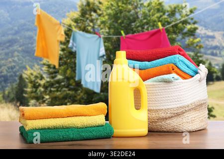 Korb mit Wäsche und Flasche Waschmittel auf Holztisch in der Nähe von Kleidung, die draußen trocknet Stockfoto