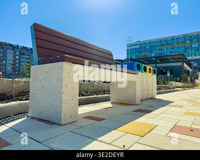 In einem Stadtpark befindet sich eine Bank aus massiven Materialien. In der Nähe befinden sich Recyclingbehälter in verschiedenen Farben. Die Sonne scheint hell am Himmel. Residenti Stockfoto