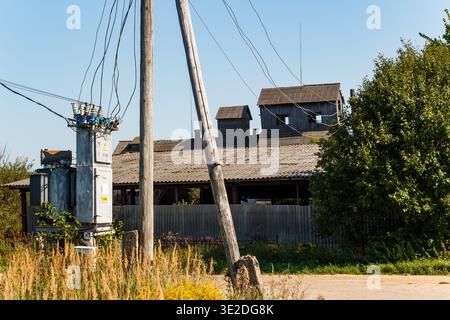 Alte ländliche Stromtransformatoren und Stangen stehen vor einem klaren Himmel, mit verwitterten Holzgebäuden und trockenem Gras in einer rustikalen Dorfkulisse Stockfoto