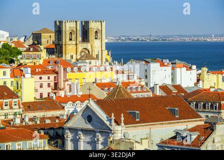 Farbenfrohe Dächer von Lissabons Alfama-Viertel mit der historischen Kathedrale Sé und einem Frachtschiff auf dem Tejo unter einem hellblauen Himmel. Stockfoto