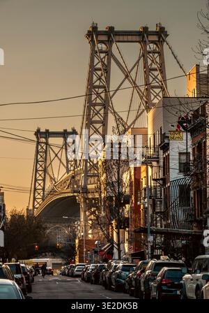Brooklyn, NY - USA - 14. März 2026 die Golden Hour-Leuchte umrahmt die Williamsburg Bridge über einer klassischen Brooklyn-Straße mit Autos, Ziegelgebäuden und urbanen Gebäuden Stockfoto