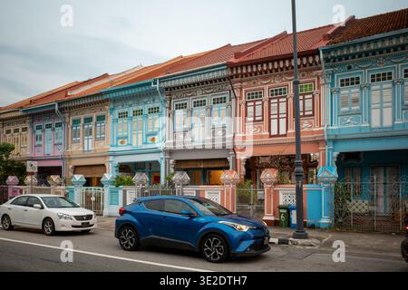 Farbenfrohe Geschäfte säumen eine ruhige Straße. Blaues Auto parkt neben einer weißen Limousine. Traditionelle Architektur mit Fliesendächern. Pastelltöne zieren jedes Gebäude Stockfoto