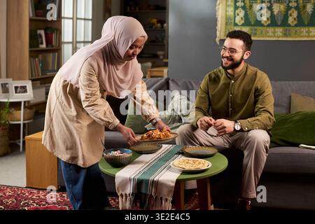 Junge muslimische Frau, die traditionelles Essen für jungen erwachsenen muslimischen Mann aus dem Nahen Osten serviert, der auf Sofa sitzt, und Paare, die gemeinsam im Wohnzimmer essen Stockfoto