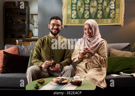 Porträt eines jungen erwachsenen muslimischen Mannes und einer jungen erwachsenen muslimischen Frau, die auf dem Sofa sitzen und lächelnd Teetassen, traditionelle Snacks und Teekanne auf dem Tisch im Wohnzimmer halten Stockfoto