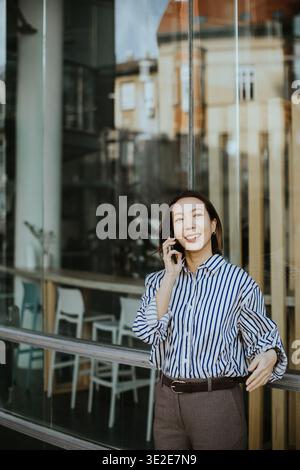 Eine Koreanerin steht vor einem modernen Gebäude. Sie lächelt, während sie am Telefon spricht. Das Sonnenlicht scheint auf sie, während sie sich mit jemandem verbindet. Stuhl Stockfoto