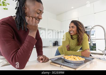 Afroamerikanischer Mann und indische Frau, die sich in Pullis auf die Küchenzeile lehnen und Kuchen bewundern Stockfoto