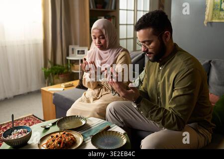 Junge Erwachsene muslimische Frau und junger erwachsener muslimischer Mann aus dem Nahen Osten sitzen auf dem Sofa und beten mit erhobenen Händen vor dem Essen, traditionelle Speisen werden auf dem Tisch im Wohnzimmer serviert Stockfoto