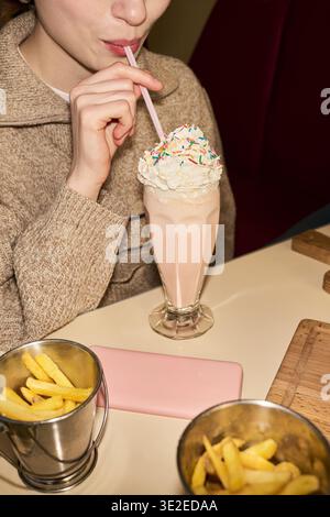 Vertikale Aufnahme einer jungen Frau in der Mitte, die Milchshake mit Schlagsahne trinkt und durch Strohhalm streut, während sie am Tisch sitzt und Pommes Frites im Vordergrund hat Stockfoto