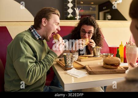 Gesellschaft von hungrigen Freunden, die am Tisch sitzen und Burger im Diner essen, beide konzentrierten sich auf Essen, Edelstahlbecher und Gewürze auf dem Tisch, drittens teilweise sichtbar Stockfoto