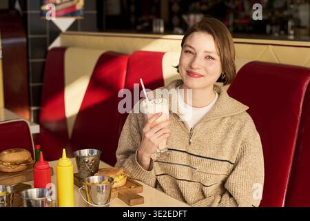 Porträt einer jungen kaukasischen Frau, die lächelt, während sie einen Milchshake mit Strohhalm hält, in einem Restaurant im Retro-Stil sitzt, Burger und Gewürze auf dem Tisch und die Kamera betrachtet Stockfoto