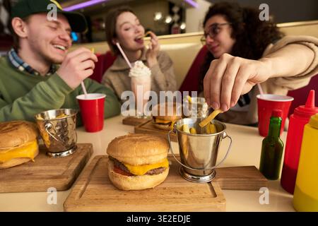 Eine Gruppe von drei Schülern sitzt am Tisch und isst Cheeseburger und Pommes, trinkt Milchshake und Limonade und genießt gemeinsam eine zwanglose Mahlzeit Stockfoto