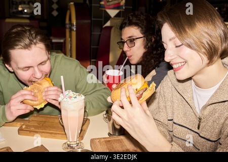Drei Z-Freunde der Generation essen Burger und trinken Milchshake am Restauranttisch, lächeln und genießen gemeinsam das Essen Stockfoto
