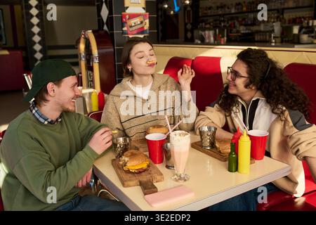 Drei junge Freunde sitzen zusammen im Retro-Diner, lächeln und lachen, während sie Burger essen und Milchshakes trinken und sich freundlich unterhalten Stockfoto
