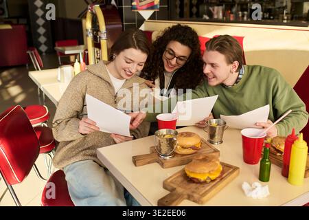 Gruppe junger Freunde, die zusammen im Diner sitzen und lächelnd Menüs, Burger und Getränke auf dem Tisch anschauen und die gemeinsame Zeit genießen Stockfoto