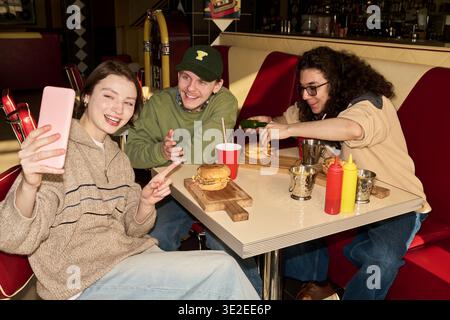 Gesellschaft von drei jungen Freunden, die am Esstisch sitzen, lächeln und Selfie mit dem Smartphone machen, während sie Burger essen und Limonade trinken Stockfoto